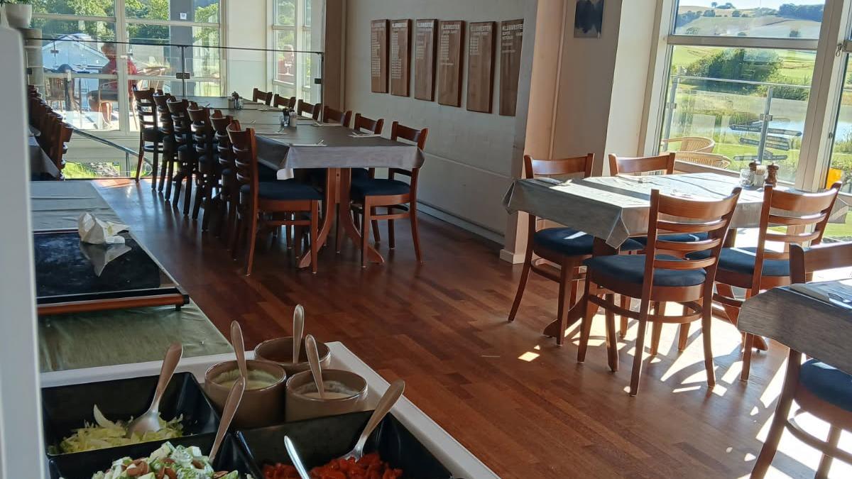 Indendørs restaurant med træborde, stole og en salatbar med forskellige ingredienser. Sollys strømmer ind gennem vinduerne.