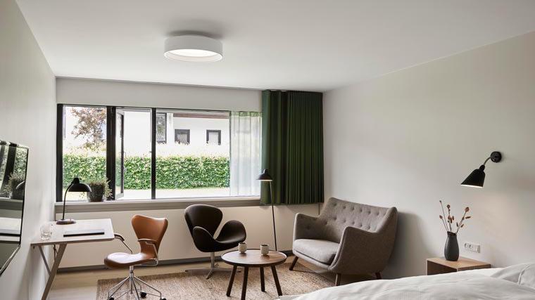 Moderne hotelværelse med vindue, sofa, lænestole og skrivebord i lys indretning.