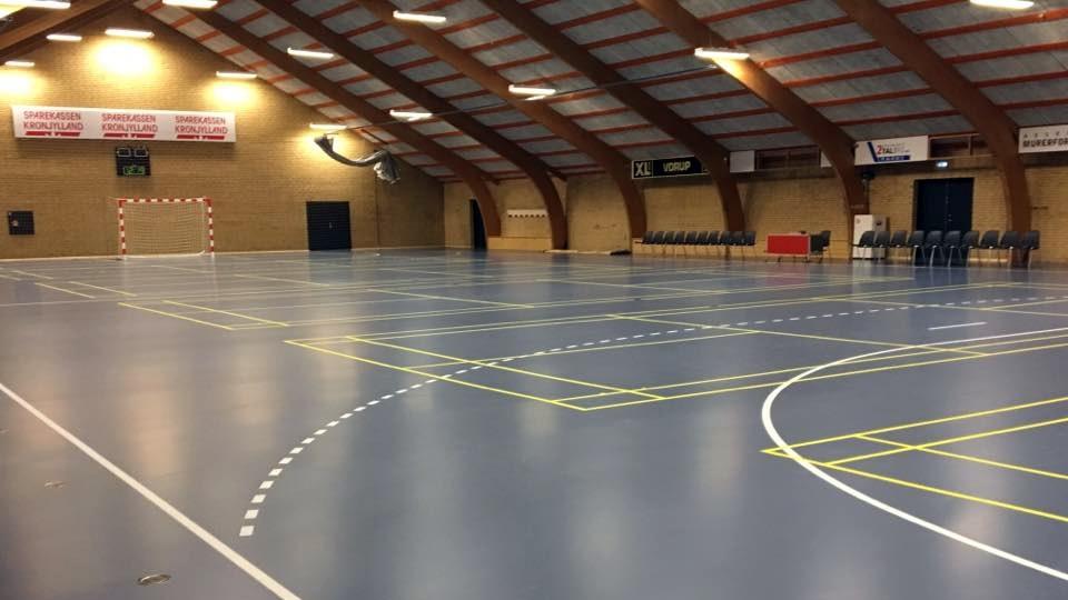 Indendørs sportshal med blåt gulv, håndboldmål og markerede baner til forskellige sportsgrene.