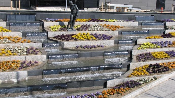 En skulptur af en mand står i en blomsterhave med farverige blomster og vandfald i baggrunden.