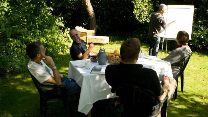 Gruppe af mennesker, der sidder omkring et bord i haven, mens en person præsenterer på en tavle.