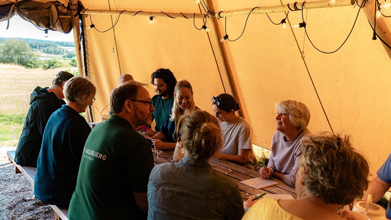 Gruppe mennesker sidder ved et langbord under en telt, der diskuterer og arbejder sammen i naturen.