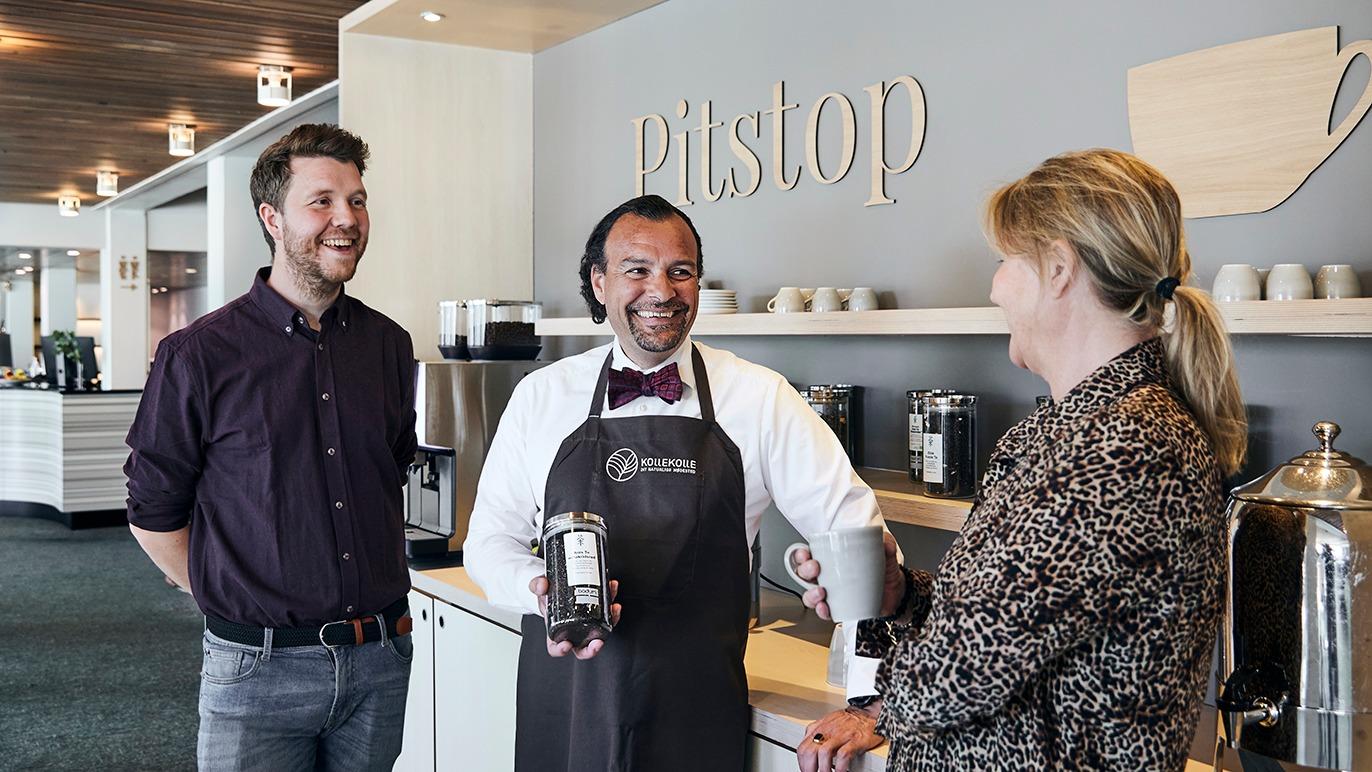Tre personer står ved en kaffebar, hvor en barista serverer kaffe til to kunder.