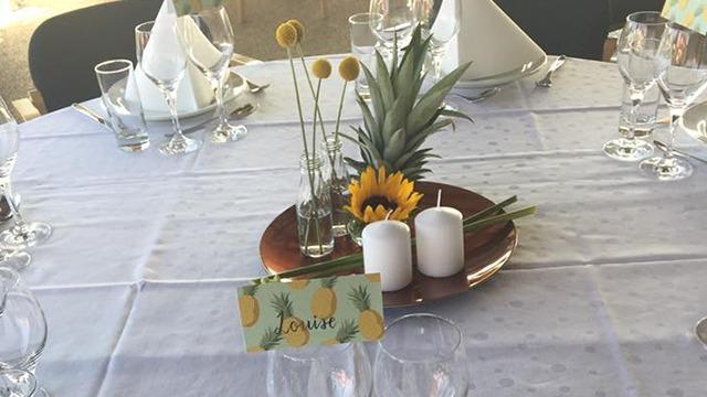Et festligt dækket bord med glas, servietter, en ananas, solblomster og stearinlys på en kobberplade.
