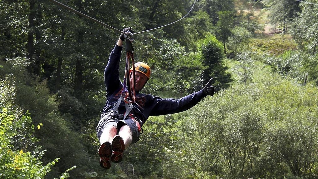 En mand svæver ned ad en zipline gennem en grøn skov med træer i baggrunden.