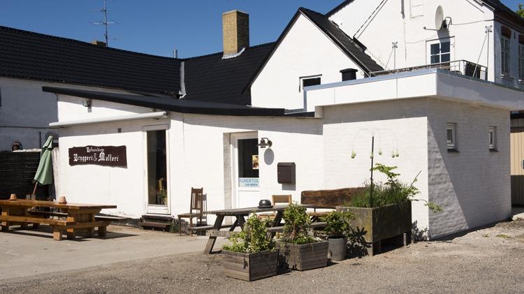 En hvid bygning med en café, udendørs siddepladser og planter i potter under en klar blå himmel.