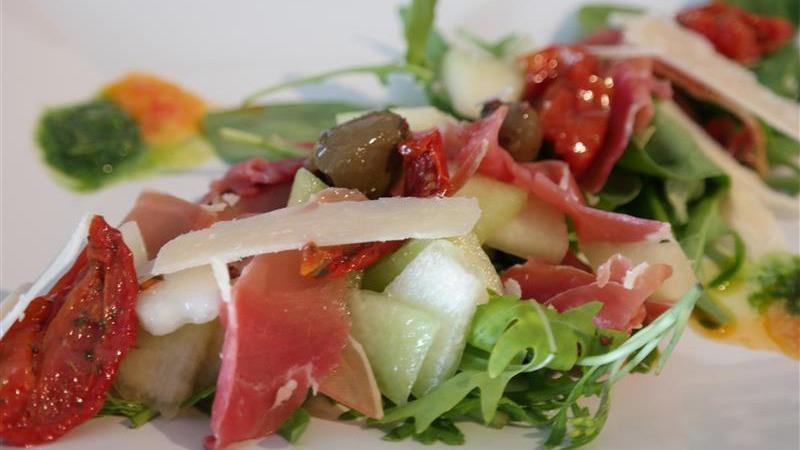 En farverig salat med skinke, soltørrede tomater, melon, oliven og parmesan på en hvid tallerken.