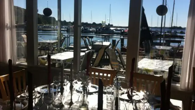 Udsigt fra en restaurant med borde og stole, der vender mod en havn med både og solskin.