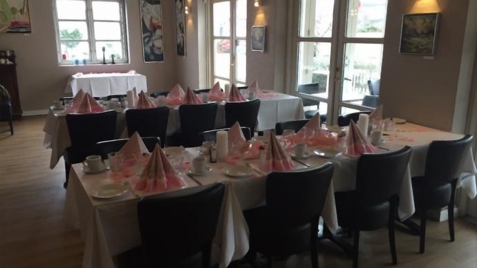 Et elegant restaurantlokale med dækkede borde, hvide duge og lyserøde servietter til en festlig begivenhed.