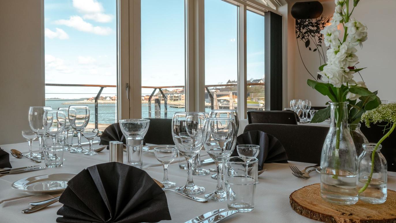 Elegant restaurantbord med glas og servietter, udsigt over vandet og sollys, skaber en indbydende atmosfære.