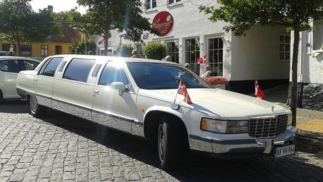 En hvid limousine med danske flag parkeret foran en bygning i en by.