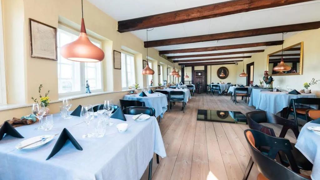 En elegant restaurant med lyse vægge, træbjælker og dækkede borde med blå duge og moderne lamper.