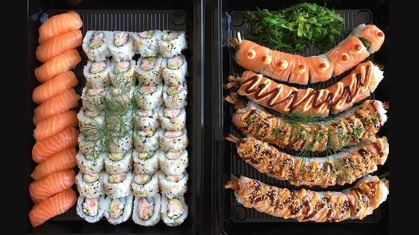 Lækre sushi-ruller og nigiri med laks, pyntet med grønt og saucer, præsenteret i en sort boks.