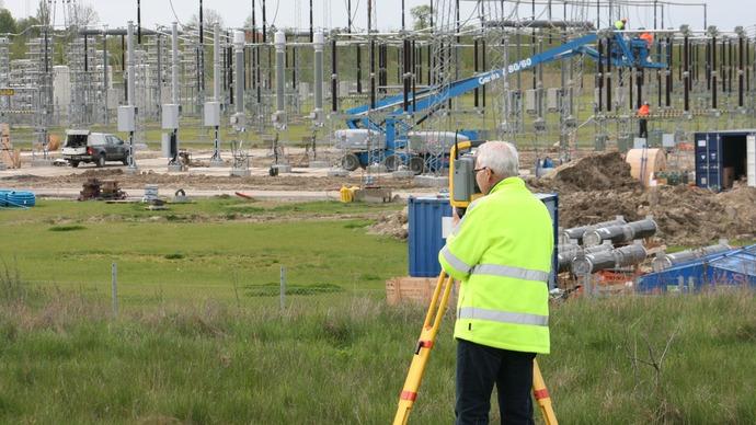En landmåler i en gul sikkerhedsvest, der arbejder ved en byggeplads for en elstation med kraner og udstyr i baggrunden.