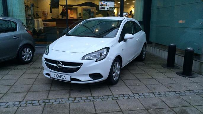 En hvid Opel Corsa parkeret foran en bygning med glasfacade og en café i baggrunden.