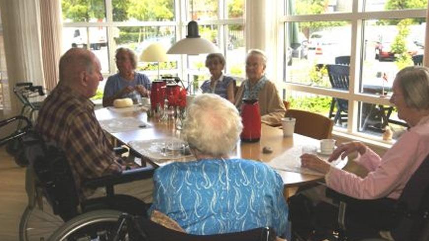 Ældre mennesker sidder omkring et bord i en lys stue, mens de nyder kaffe og samtale.