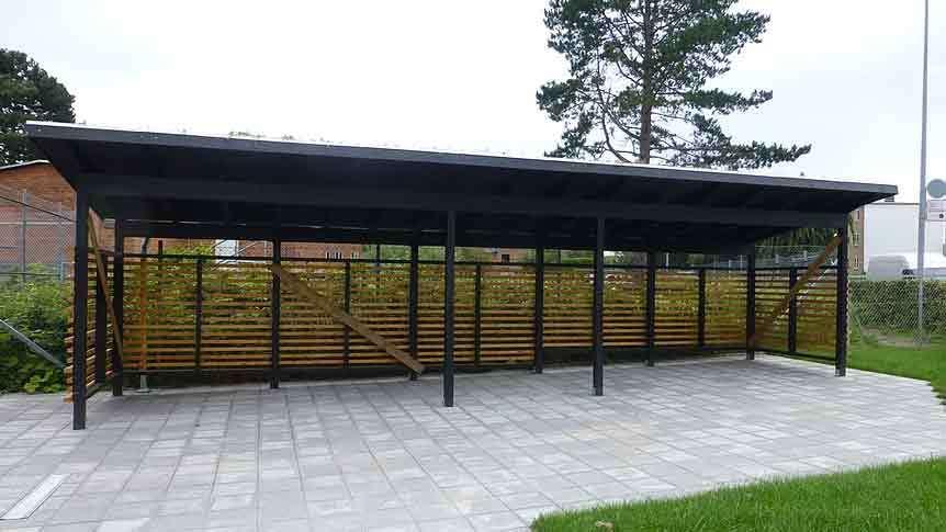 En stor carport med sort tag og trævægge, placeret på flisebelagt område med græs i forgrunden.