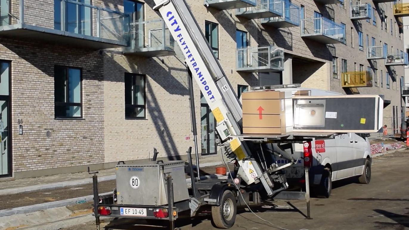 En kran løfter en stor kasse fra en trailer ved en nybygget lejlighedsbygning.