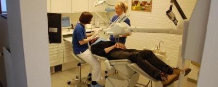 To personer i blå skjorter arbejder i en tandklinik, mens en patient ligger i tandlægestolen.