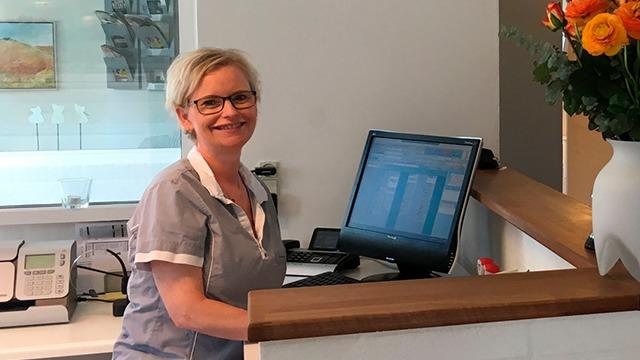 En smilende receptionist i en moderne klinik med en computer og blomster på skranken.