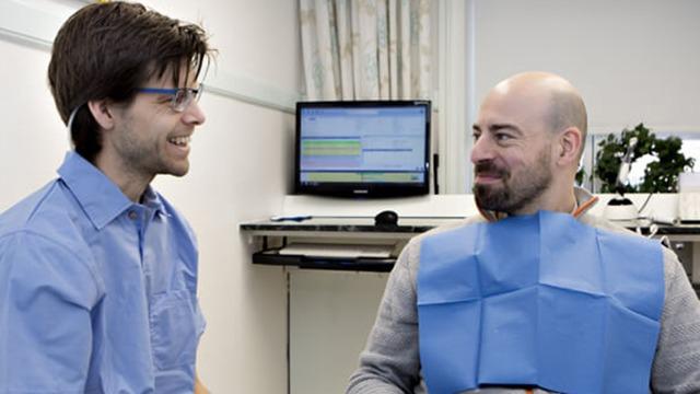 To mænd i en tandlægeklinik, hvor den ene smiler og den anden sidder med en blå beskyttelsesforklæde.