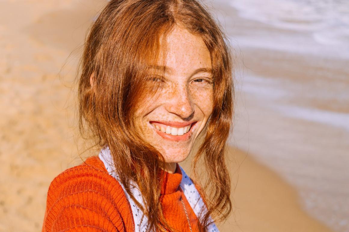 En smilende kvinde med rødt hår og fregner står på stranden ved havet. Hun bærer en rød trøje og ser glad ud.