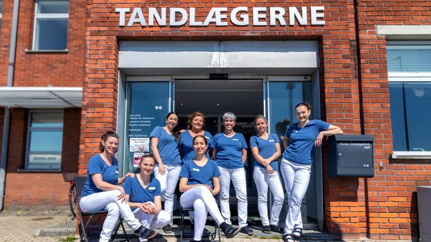 En gruppe tandlæger og personale står foran tandklinikken, iført blå uniformer og smilende.