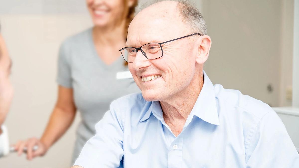 En glad ældre mand med briller smiler, mens han taler med en sygeplejerske i en klinik.