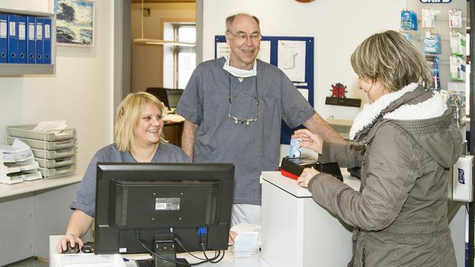 To ansatte i en klinik står ved skranken, mens en patient betaler for behandling. Der er en computer på bordet.