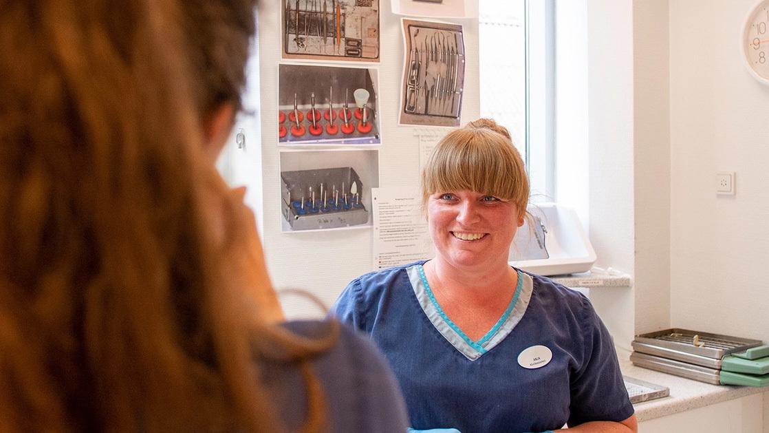 En smilende kvinde i arbejdstøj står ved en skranke og taler med en kunde i en lys klinik.