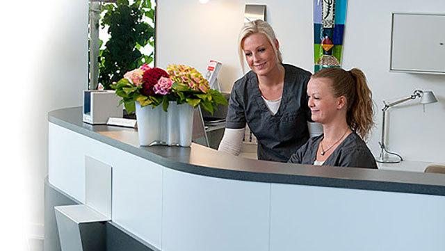 To kvinder i arbejdstøj står ved en reception med blomsterarrangementer og smiler til hinanden.