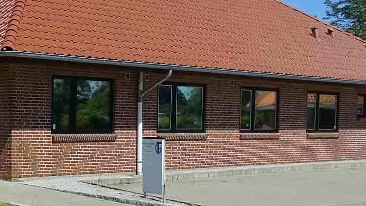 En murstensbygning med rødt tag og flere vinduer, beliggende i et solrigt område.