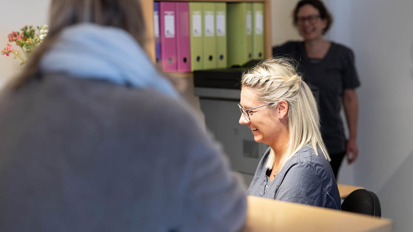 En smilende kvinde ved receptionen med mapper i baggrunden og en anden kvinde i grå tøj i fokus.