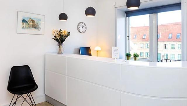 Moderne reception med hvidt skrivebord, planter, vægur og kunstværk, beliggende ved vinduet med udsigt.