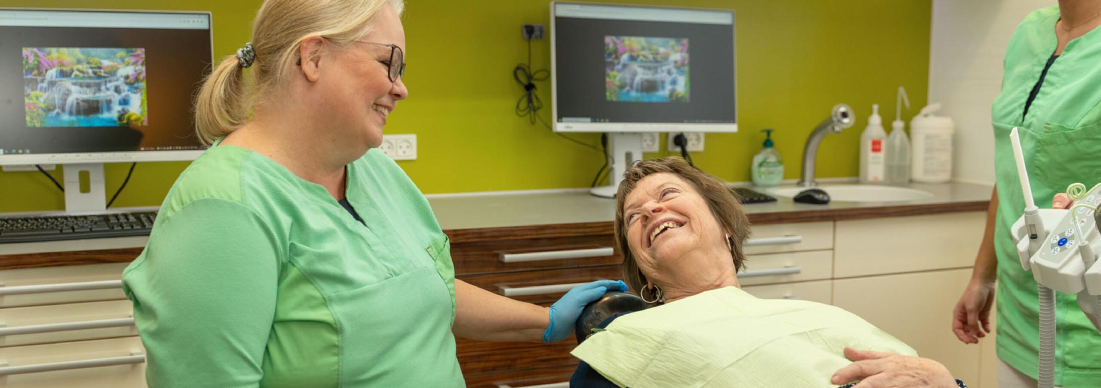 En tandlæge og en patient smiler og griner sammen i en moderne tandklinik med computere i baggrunden.