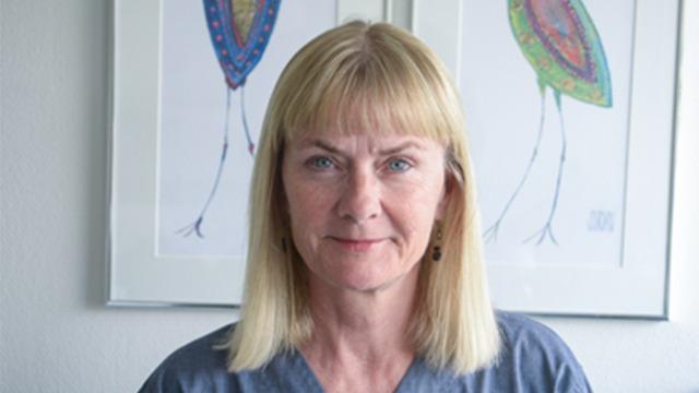 Kvinde med blond hår står foran farverige fugleillustrationer på væggen.