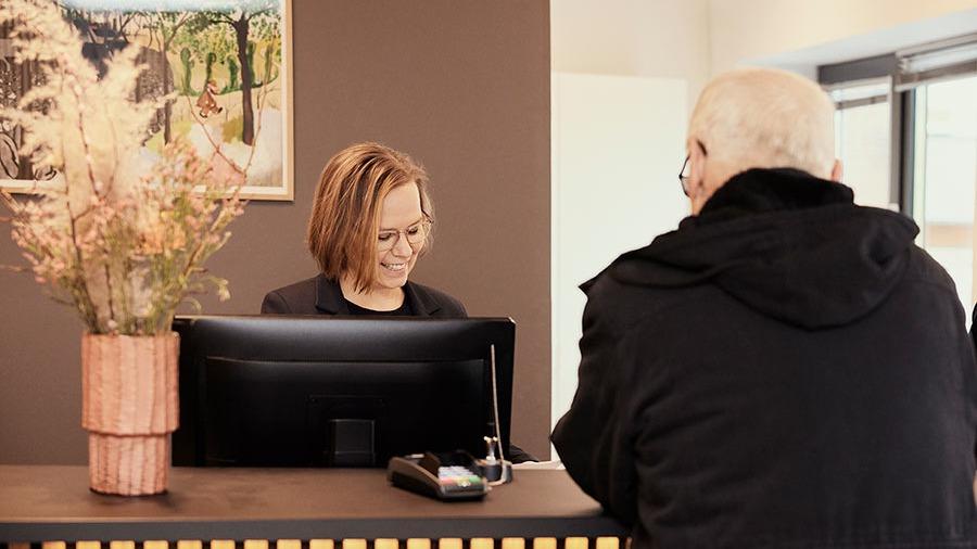 En receptionist smiler til en gæst ved skranken i en moderne reception med blomsterdekoration.