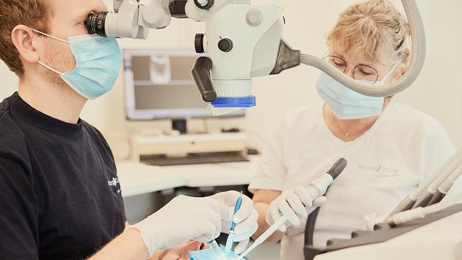 En tandlæge og en assistent arbejder sammen under en tandbehandling med mikroskop. Begge bærer ansigtsmasker.