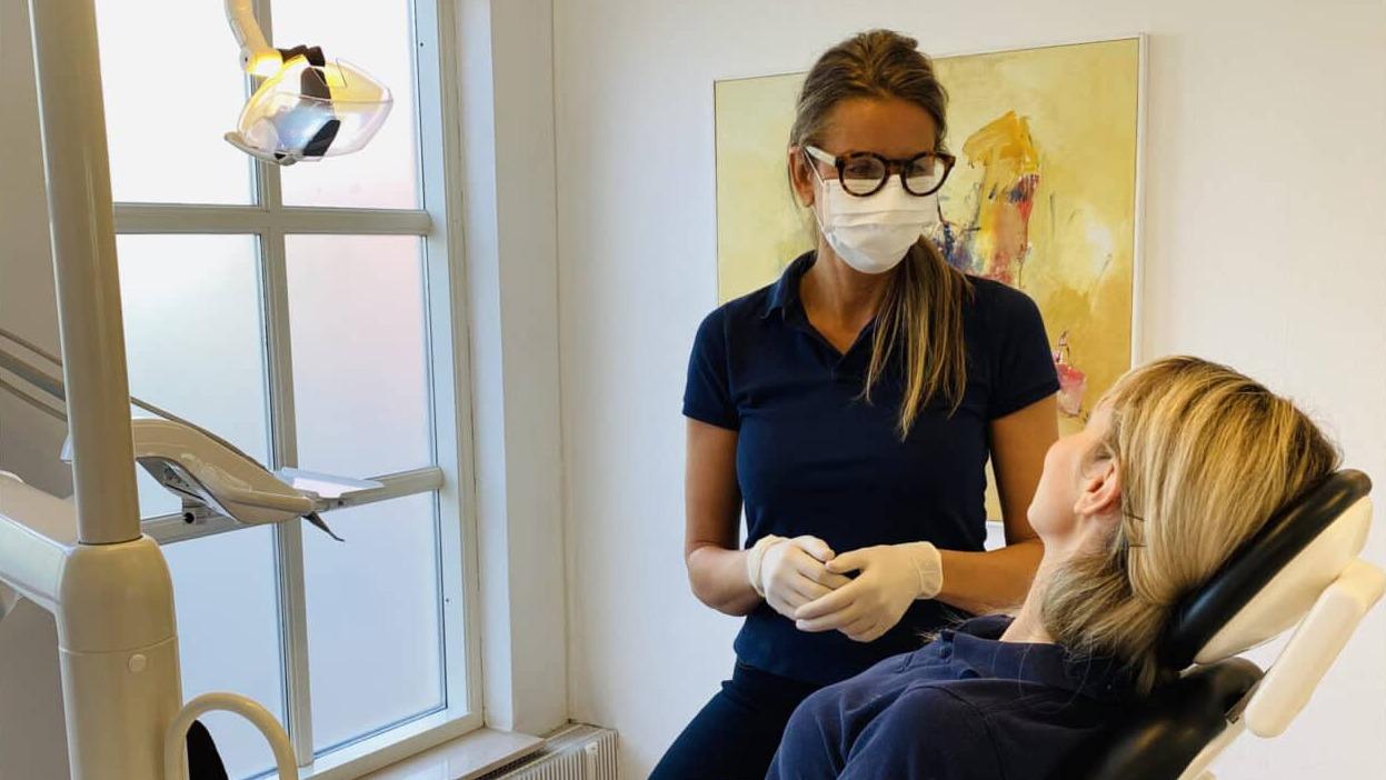 En tandlæge i beskyttelsesudstyr står ved en patient i en tandlægestol i et lyst behandlingsrum.