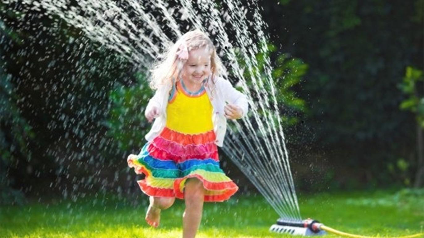 En glad pige i farverig kjole løber gennem vandsprinkler i haven på en solrig dag.