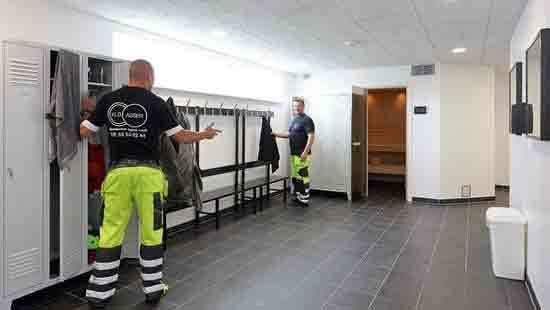 Toiletter og omklædningsrum med ansatte i arbejdstøj, der hænger tøj op i skabe og snakker sammen.