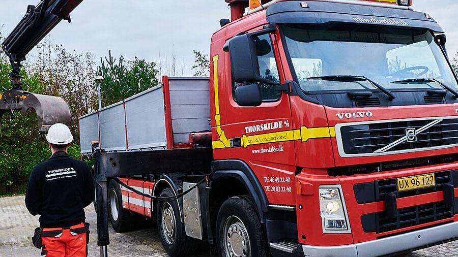 En rød Volvo lastbil med en kran, hvor en mand i sikkerhedshjelm overvåger arbejdet.