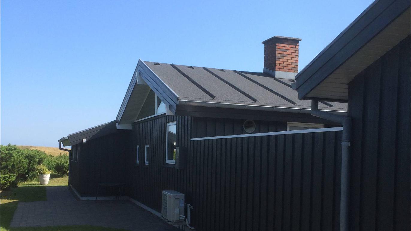 Moderne sort hus med skråtag og skorsten, beliggende i et grønt område under en klar blå himmel.