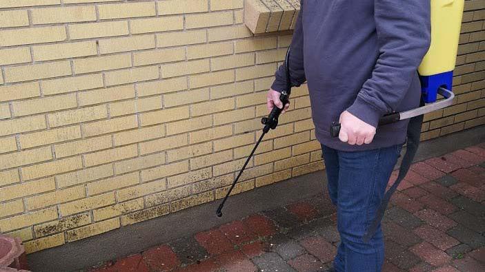 En person med en rygsprøjte står ved en mur og sprøjter mod snavs på fliserne.