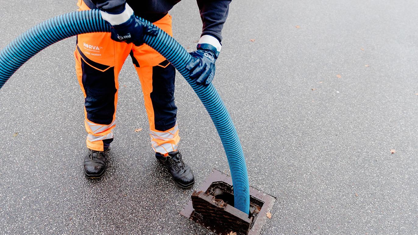 En mand i orange arbejdstøj tilslutter en blå slange til en brønd på en asfaltvej.