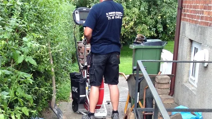 En mand står på en maskine i en have, omgivet af buske og en affaldscontainer.