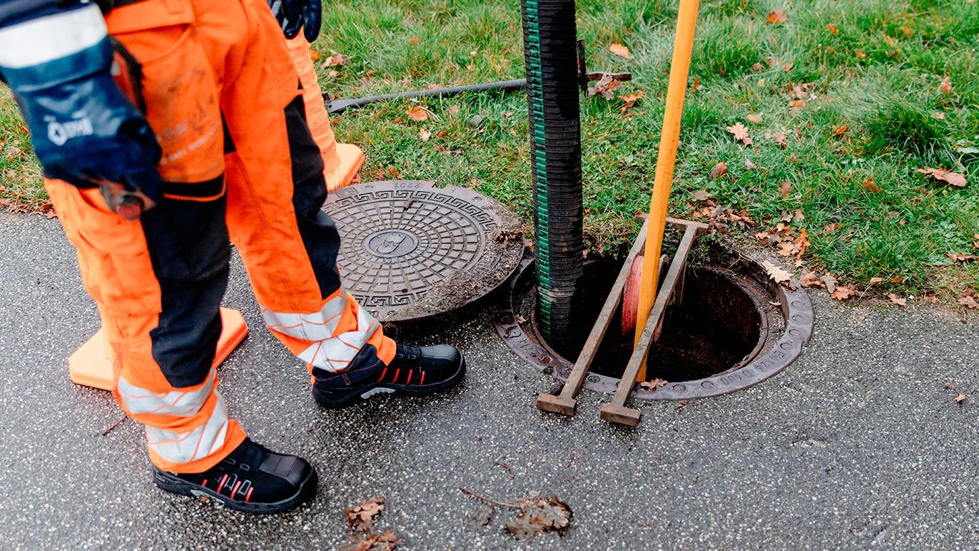 En arbejdstager i orange arbejdstøj står ved en åben brønd med værktøj i hånden.