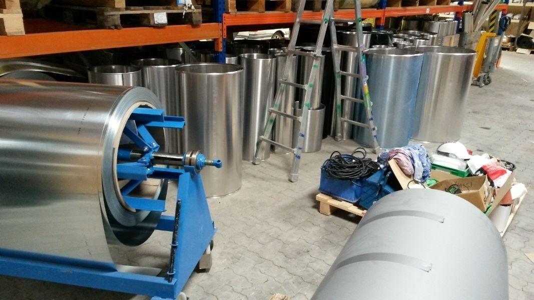 Lager med metalruller og en blå maskine, samt en stige og diverse værktøj i baggrunden.