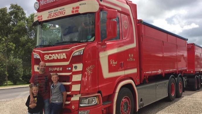 En rød Scania lastbil med to børn og en voksen stående foran, på en grusvej med træer i baggrunden.