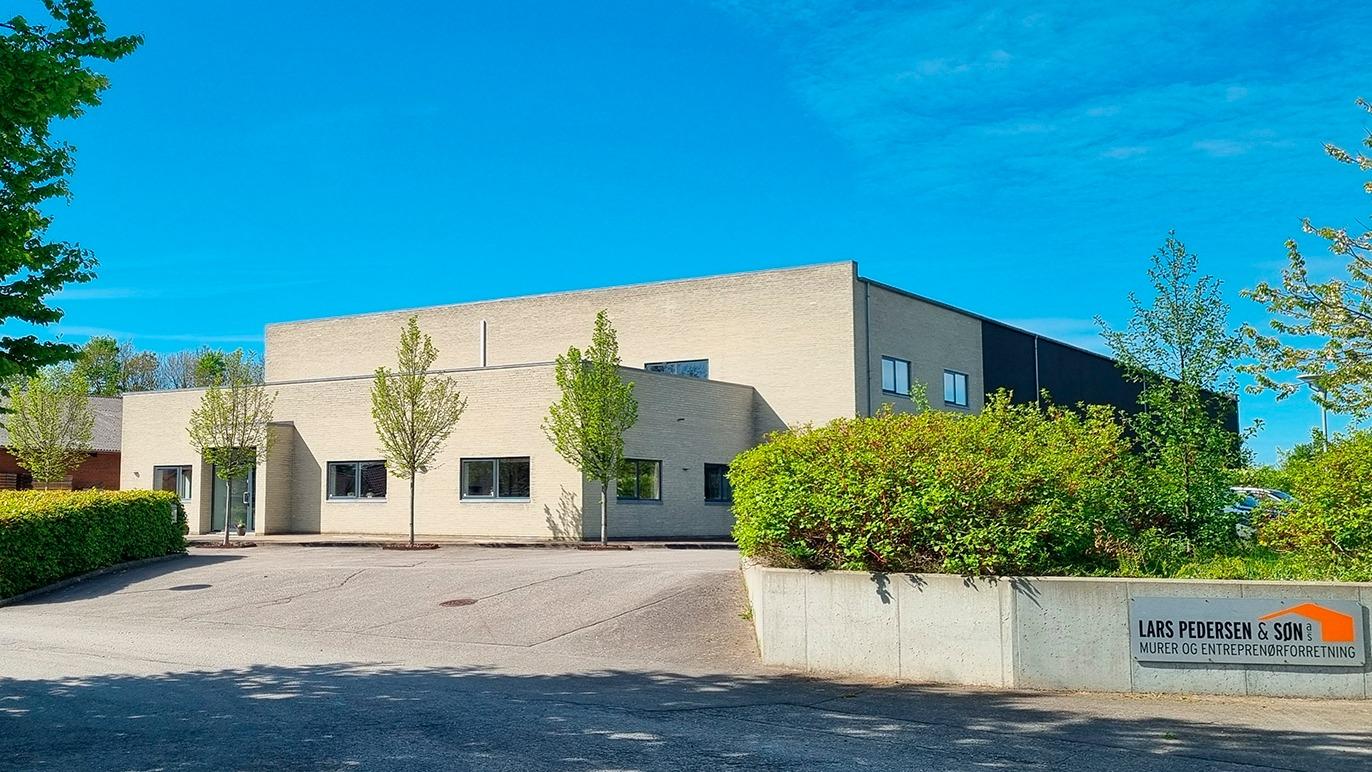 Moderne bygning med grå facade, omgivet af træer og buske, beliggende på en asfalteret vej.